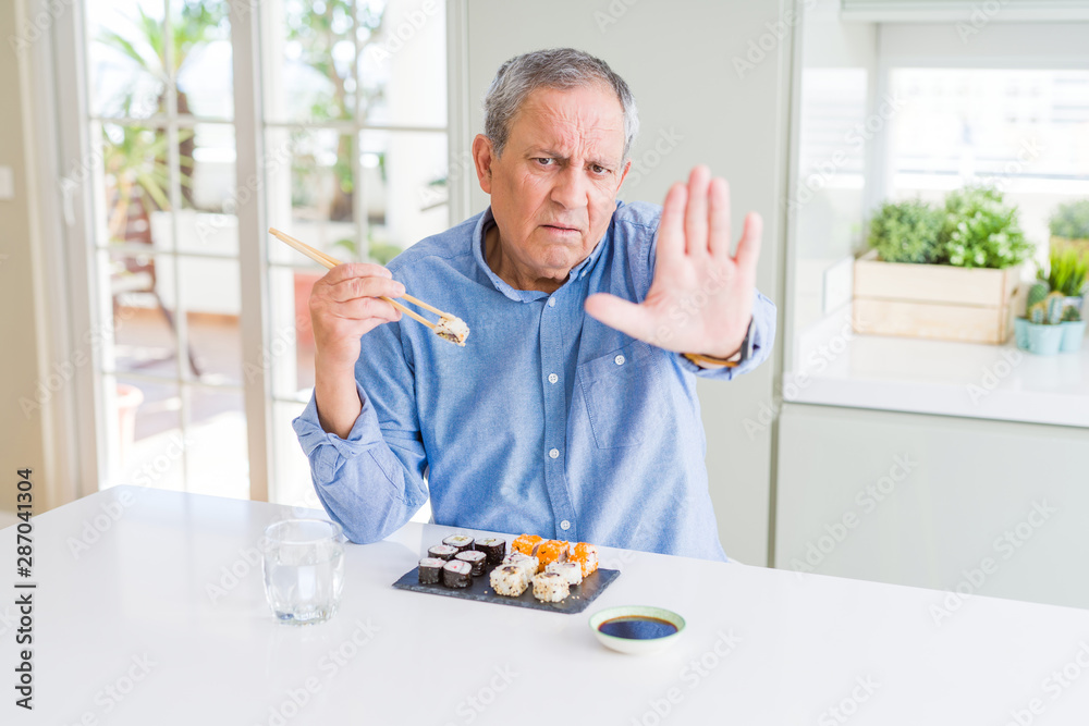 Handsome senior man eating take away sushi using chopsticks at home with open hand doing stop sign with serious and confident expression, defense gesture Handsome senior man eating take away sushi using chopsticks at home with open hand doing stop sign with serious and confident expression, defense gesture