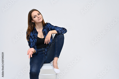 Portrait of a cute brunette girl, a young woman with beautiful curly hair in a blue shirt and jeans, is sitting on a chair on a white background. Smiling, talking with emotions, showing hands.