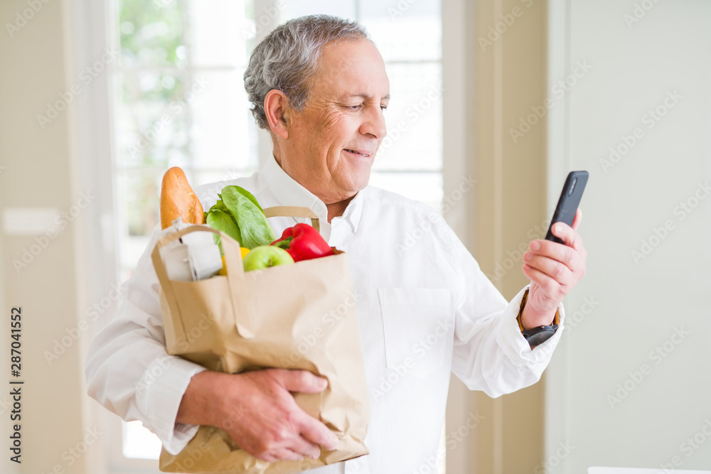 Handsome senior man holding paper bag full of fresh groceries and looking at smartphone smiling