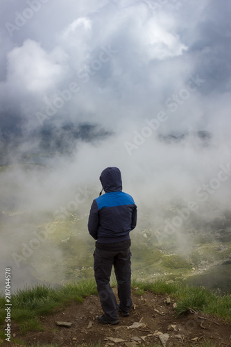 Hiker with blue jacket and hood over head standing at the edge of Lake peak and looking at seven Rila lakes through thick fog