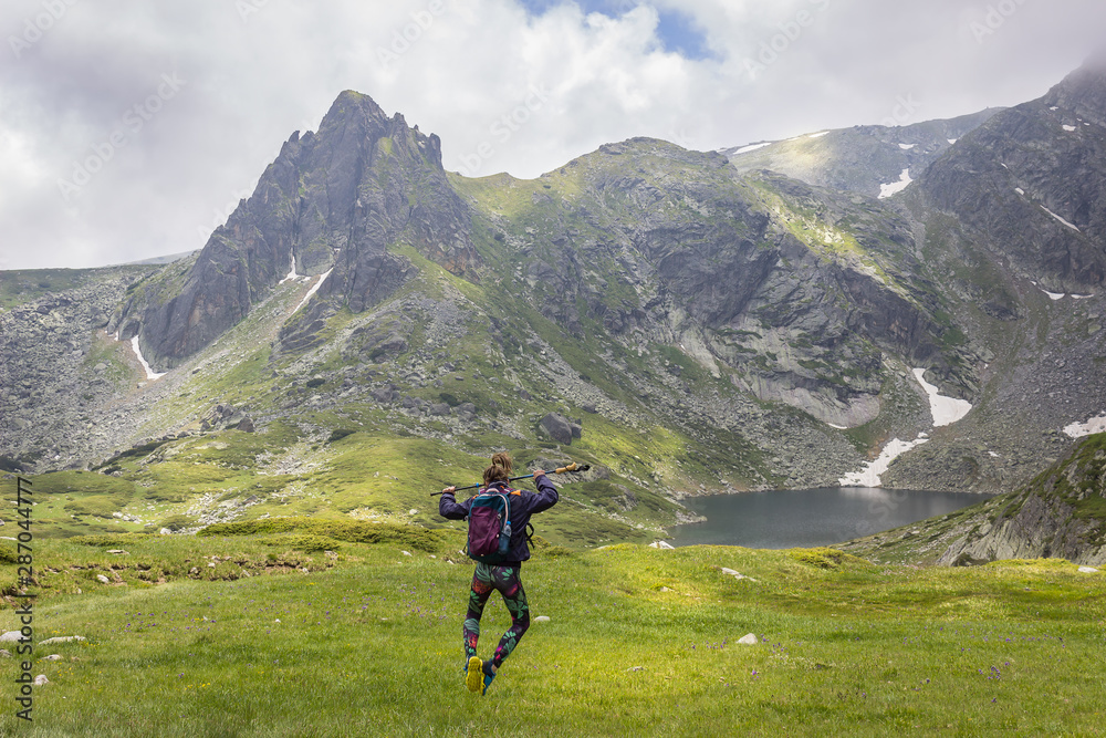 Sporty girl with backpack, tights and hiking stick jumping in front of the Twin glacial lake on Rila mountain and epic pointy summit Sporty girl with backpack, tights and hiking stick jumping in front of the Twin glacial lake on Rila mountain and epic pointy summit