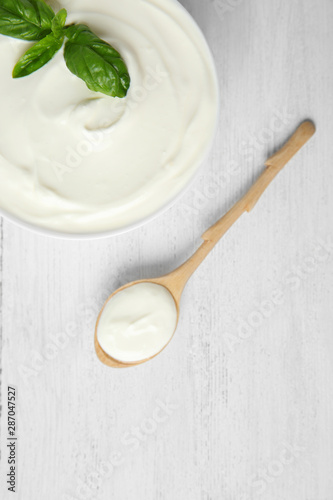 Bowl of fresh sour cream with basil and spoon on white wooden table, flat lay