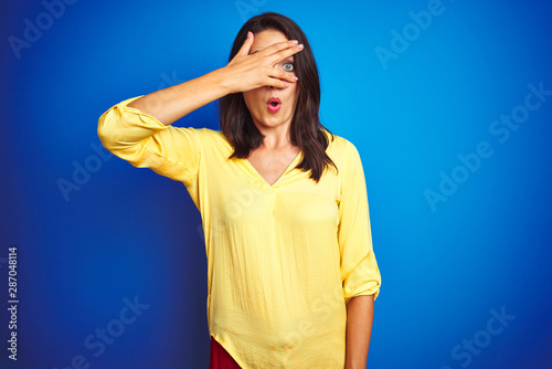 Young beautiful woman wearing yellow t-shirt standing over blue isolated background peeking in shock covering face and eyes with hand, looking through fingers with embarrassed expression. © Krakenimages.com