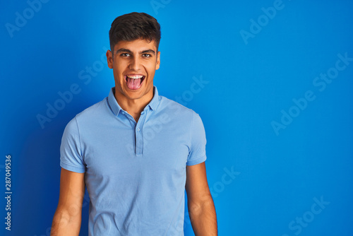 Young indian man wearing casual polo standing over isolated blue background sticking tongue out happy with funny expression. Emotion concept.
