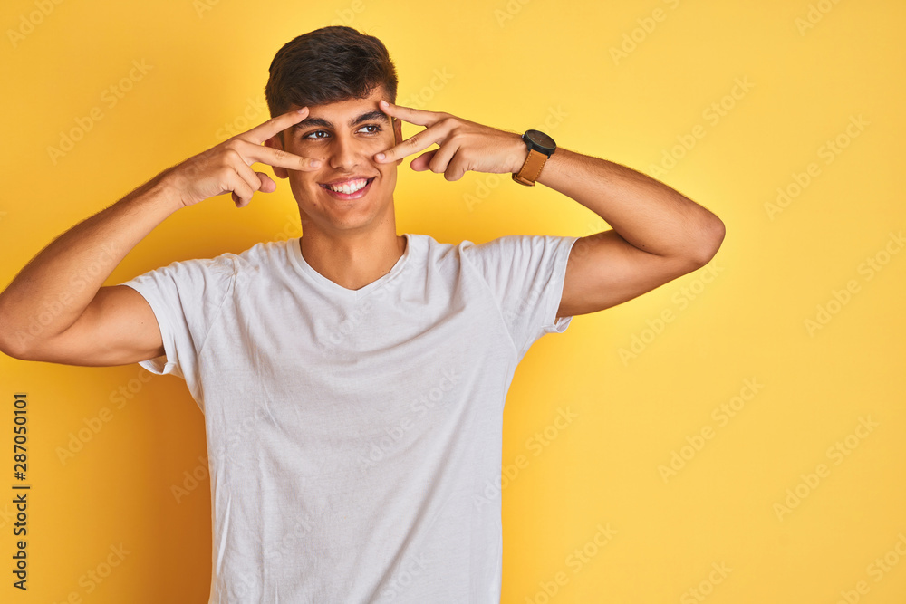 Young indian man wearing white t-shirt standing over isolated yellow background Doing peace symbol with fingers over face, smiling cheerful showing victory