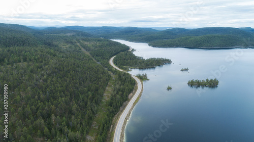 Aerial view flying over old patched two lane forest road. Green trees and lake. Shot with drone quad copter birds eye view perspective from above. 