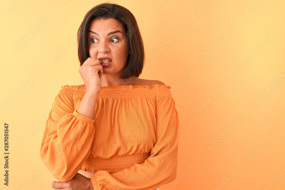 Young beautiful woman wearing casual t-shirt standing over isolated orange background looking stressed and nervous with hands on mouth biting nails. Anxiety problem. Young beautiful woman wearing casual t-shirt standing over isolated orange background looking stressed and nervous with hands on mouth biting nails. Anxiety problem.