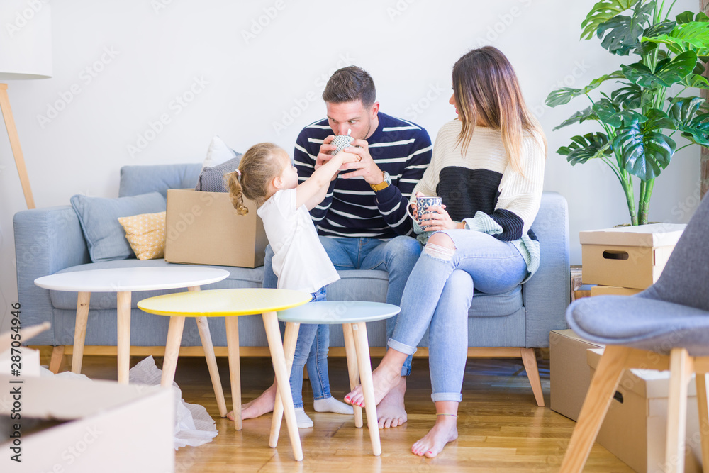 Beautiful family, parents sitting on the sofa drinking coffee looking his kid playing at new home around cardboard boxes Beautiful family, parents sitting on the sofa drinking coffee looking his kid playing at new home around cardboard boxes