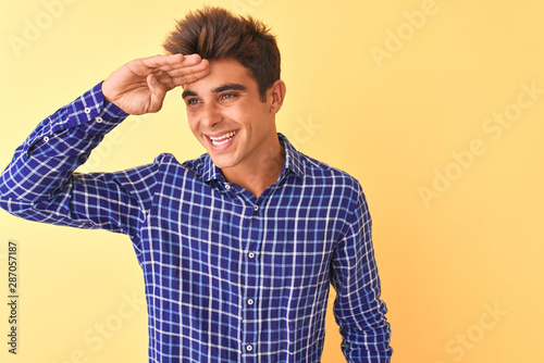 Young handsome man wearing casual shirt standing over isolated yellow background very happy and smiling looking far away with hand over head. Searching concept.