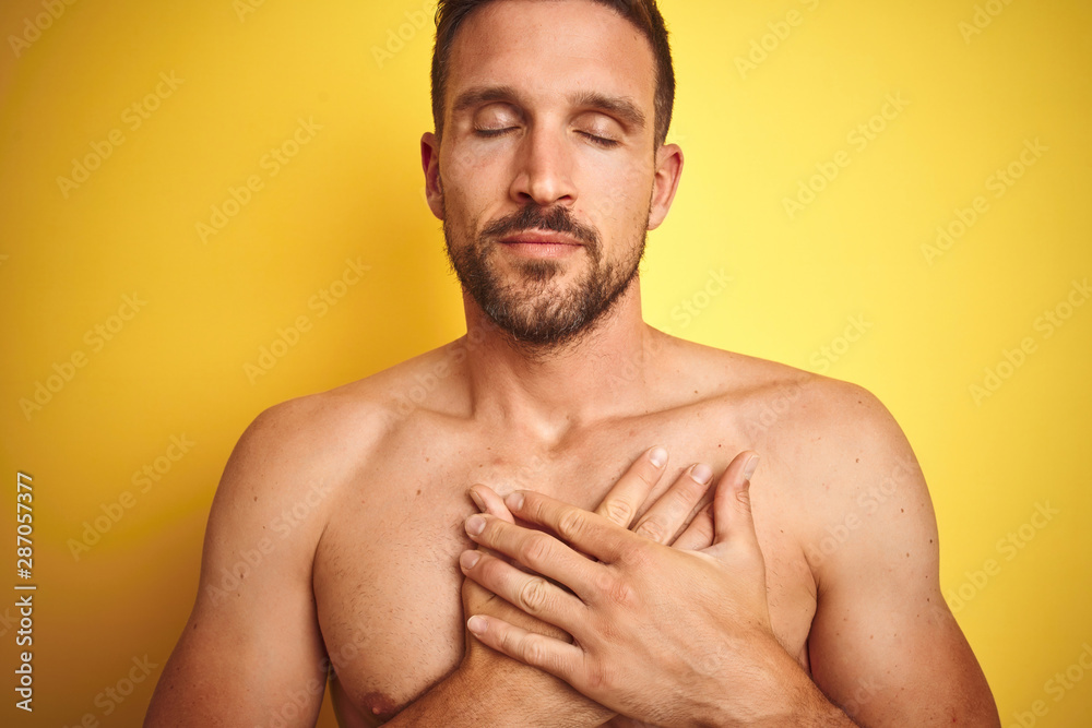 Sexy young shirtless man showing nude fitness chest over yellow isolated background smiling with hands on chest with closed eyes and grateful gesture on face. Health concept. Sexy young shirtless man showing nude fitness chest over yellow isolated background smiling with hands on chest with closed eyes and grateful gesture on face. Health concept.