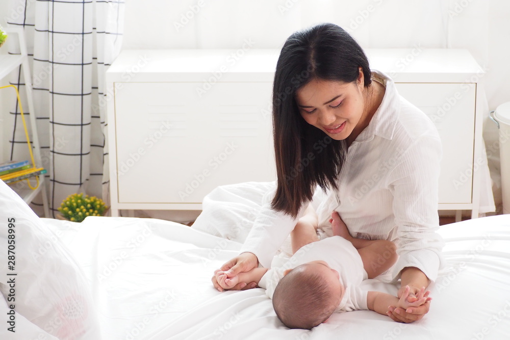 Beautiful Asian mother looking at and playing with her cute baby lying on bed in the bedroom in white and warm tone. Happy motherhood, unconditional love, lifestyle of family concept Beautiful Asian mother looking at and playing with her cute baby lying on bed in the bedroom in white and warm tone. Happy motherhood, unconditional love, lifestyle of family concept