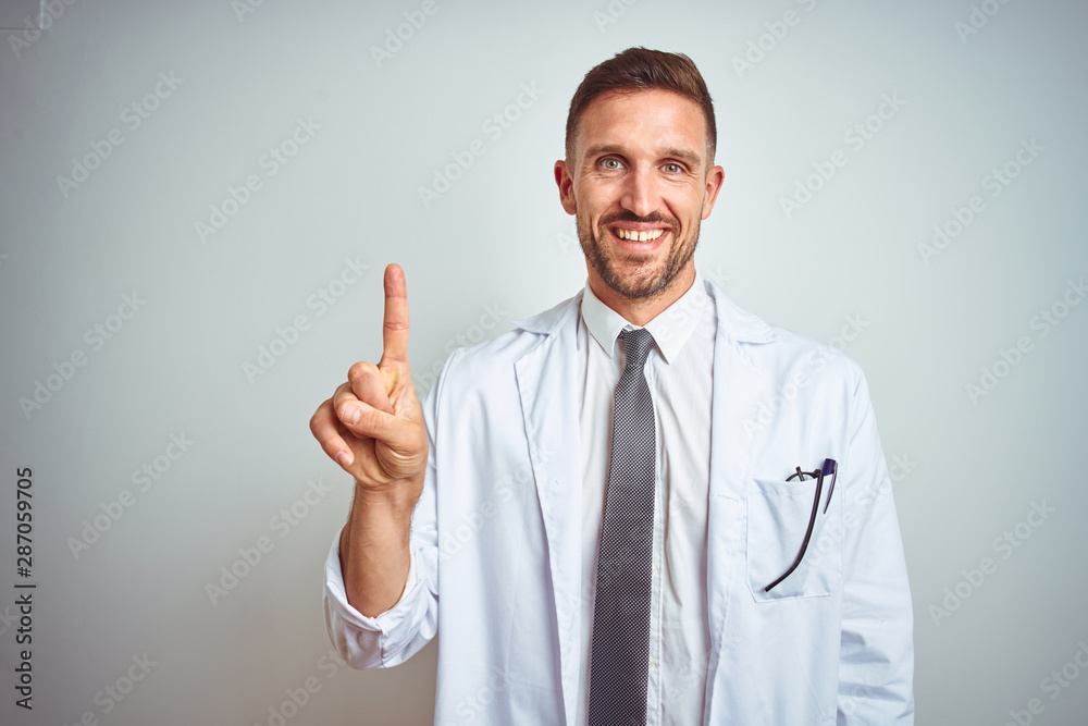Young handsome doctor man wearing white profressional coat over isolated background showing and pointing up with finger number one while smiling confident and happy. Young handsome doctor man wearing white profressional coat over isolated background showing and pointing up with finger number one while smiling confident and happy.