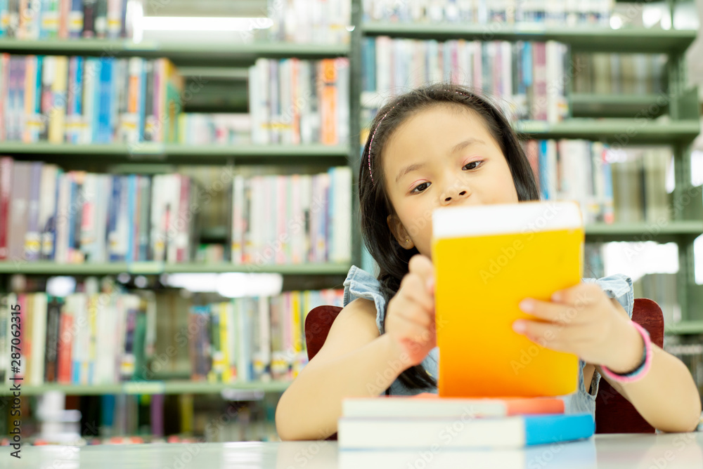 Portrait of Smiling cute Asia little Girl sitting in library at school looking to a book , education learning knowledge growth back to school concept Portrait of Smiling cute Asia little Girl sitting in library at school looking to a book , education learning knowledge growth back to school concept