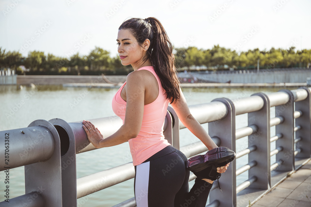 Latin woman does muscle stretching in front of a lake and dressed in pink top and black pants.
