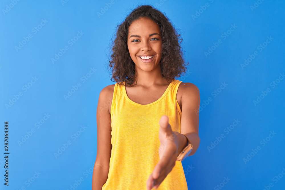 Young brazilian woman wearing yellow t-shirt standing over isolated blue background smiling friendly offering handshake as greeting and welcoming. Successful business. Young brazilian woman wearing yellow t-shirt standing over isolated blue background smiling friendly offering handshake as greeting and welcoming. Successful business.