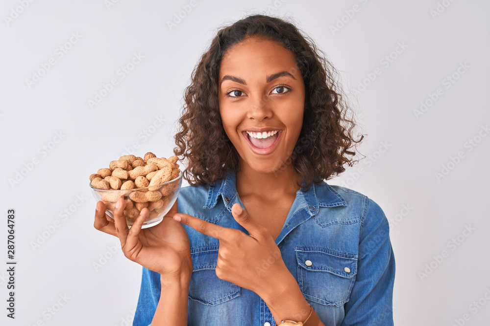 Young brazilian woman holding bowl with peanuts standing over isolated white background very happy pointing with hand and finger Young brazilian woman holding bowl with peanuts standing over isolated white background very happy pointing with hand and finger