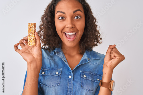 Young brazilian woman eating granola bar standing over isolated white background screaming proud and celebrating victory and success very excited, cheering emotion