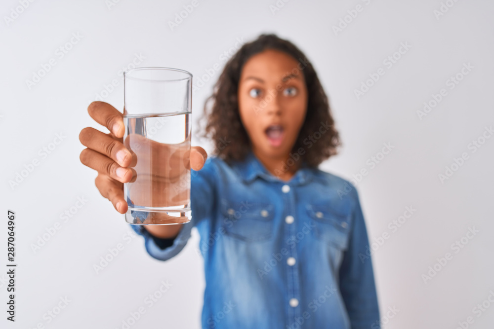 Young brazilian woman holding glass of water standing over isolated white background scared in shock with a surprise face, afraid and excited with fear expression Young brazilian woman holding glass of water standing over isolated white background scared in shock with a surprise face, afraid and excited with fear expression