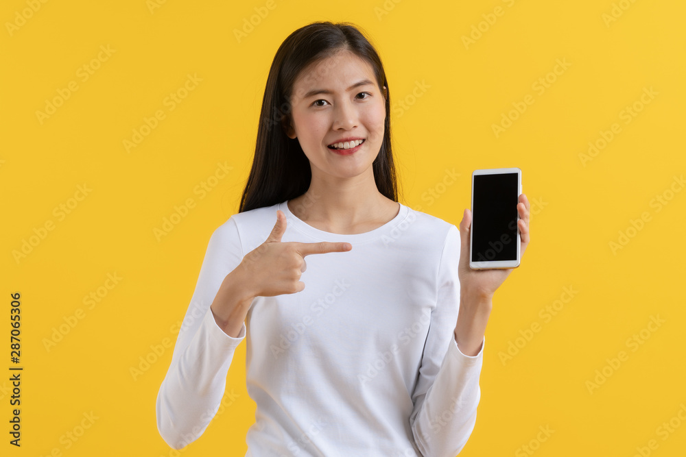 attractive asian teenager in white casual shirt pointing finger to blank mobile screen and advertising isolated on yellow background in studio. attractive asian teenager in white casual shirt pointing finger to blank mobile screen and advertising isolated on yellow background in studio.