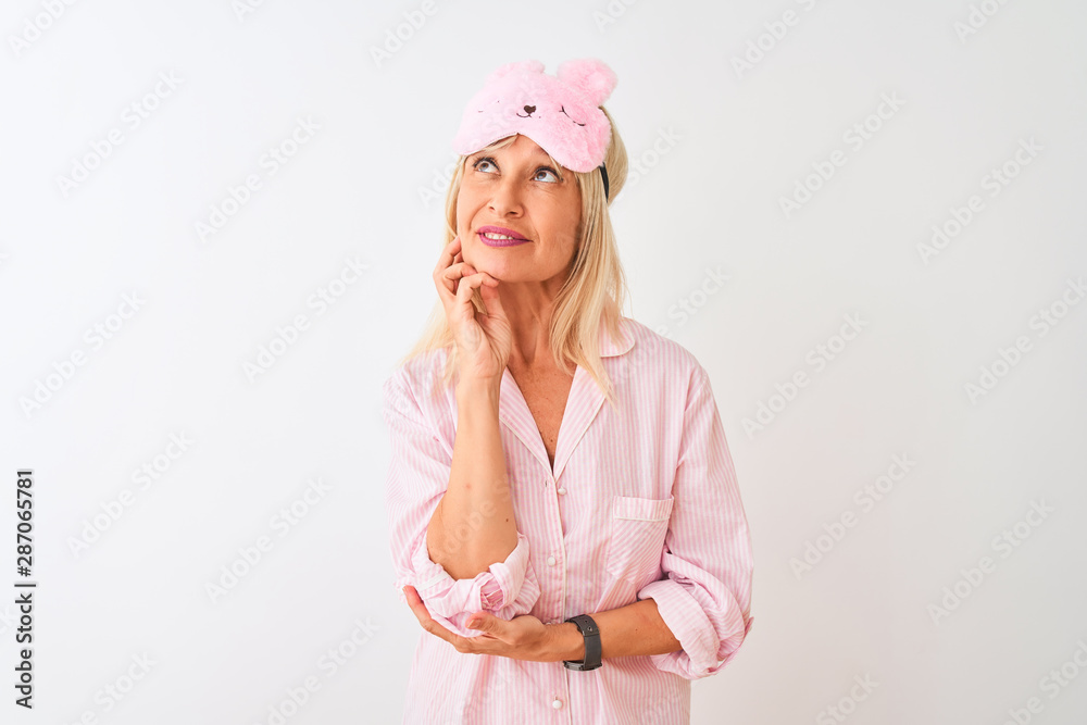 Middle age woman wearing sleep mask and pajama over isolated white background with hand on chin thinking about question, pensive expression. Smiling with thoughtful face. Doubt concept. Middle age woman wearing sleep mask and pajama over isolated white background with hand on chin thinking about question, pensive expression. Smiling with thoughtful face. Doubt concept.