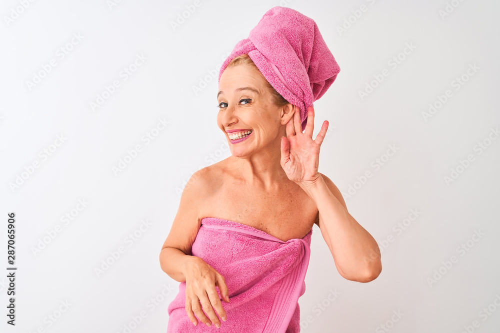 Middle age woman wearing shower towel after bath standing over isolated white background smiling with hand over ear listening an hearing to rumor or gossip. Deafness concept.