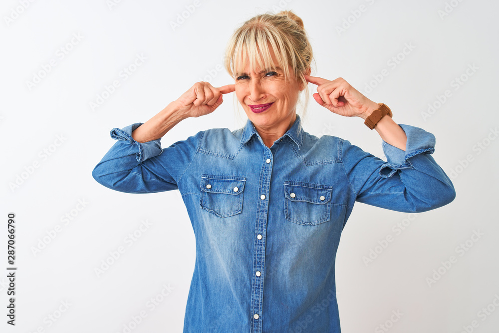 Middle age woman wearing casual denim shirt standing over isolated white background covering ears with fingers with annoyed expression for the noise of loud music. Deaf concept.