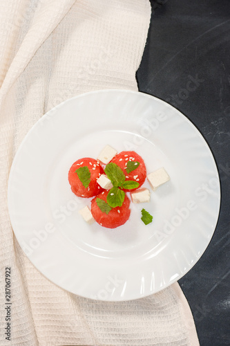 Salad of watermelon balls, feta cheese and mint, sprinkled with sesame seeds in a white plate on a black background.