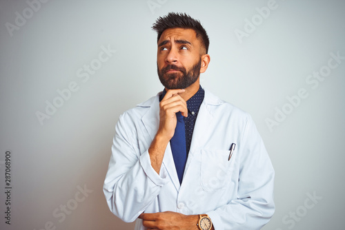 Young indian doctor man standing over isolated white background with hand on chin thinking about question, pensive expression. Smiling with thoughtful face. Doubt concept.