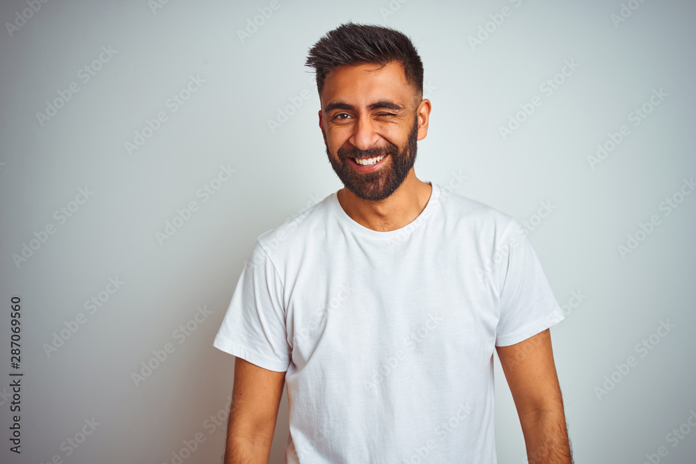 Young indian man wearing t-shirt standing over isolated white background winking looking at the camera with sexy expression, cheerful and happy face. Young indian man wearing t-shirt standing over isolated white background winking looking at the camera with sexy expression, cheerful and happy face.