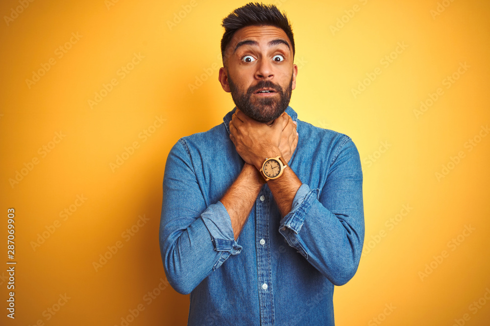 Young indian man wearing denim shirt standing over isolated yellow background shouting and suffocate because painful strangle. Health problem. Asphyxiate and suicide concept.