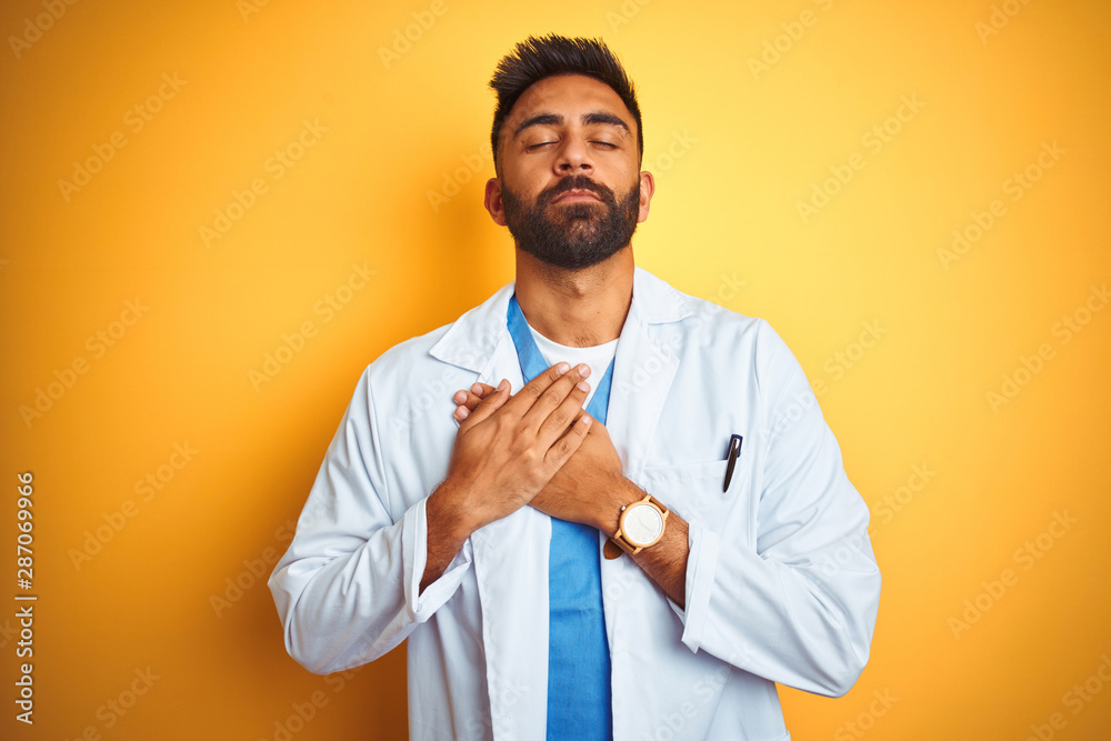 Young indian doctor man standing over isolated yellow background smiling with hands on chest with closed eyes and grateful gesture on face. Health concept. Young indian doctor man standing over isolated yellow background smiling with hands on chest with closed eyes and grateful gesture on face. Health concept.