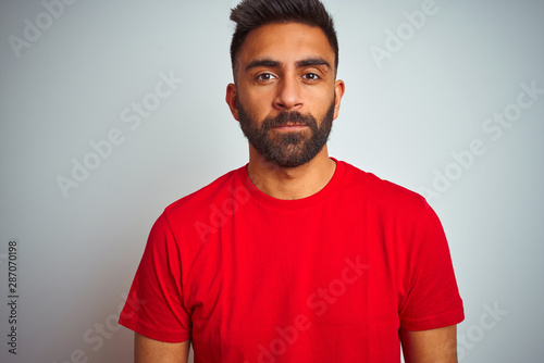 Young indian man wearing red t-shirt over isolated white background with serious expression on face. Simple and natural looking at the camera.