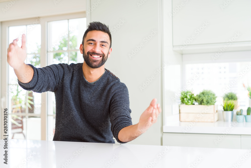 Handsome hispanic man wearing casual sweater at home looking at the camera smiling with open arms for hug. Cheerful expression embracing happiness.