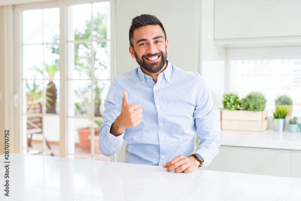 Handsome hispanic business man doing happy thumbs up gesture with hand. Approving expression looking at the camera showing success.