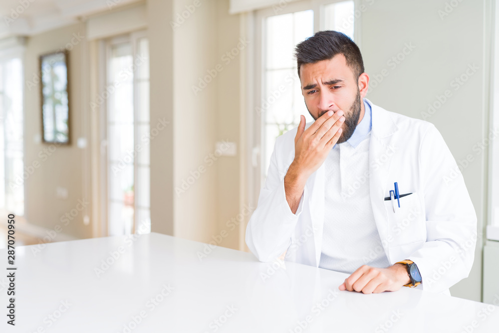 Handsome hispanic doctor or therapist man wearing medical coat at the clinic bored yawning tired covering mouth with hand. Restless and sleepiness.