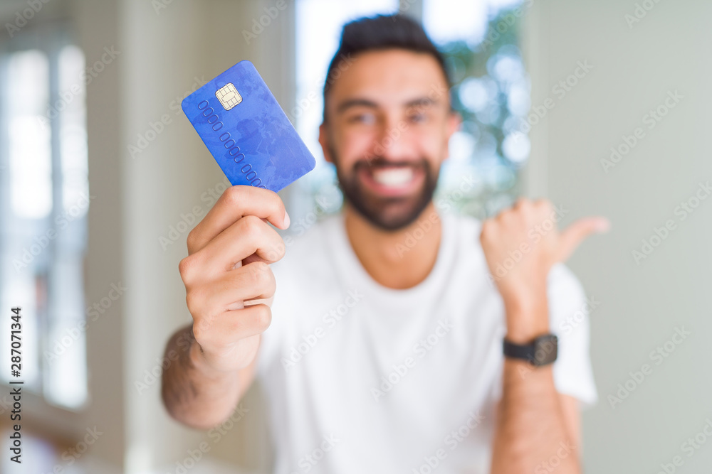 Handsome hispanic man holding credit card pointing and showing with thumb up to the side with happy face smiling Handsome hispanic man holding credit card pointing and showing with thumb up to the side with happy face smiling