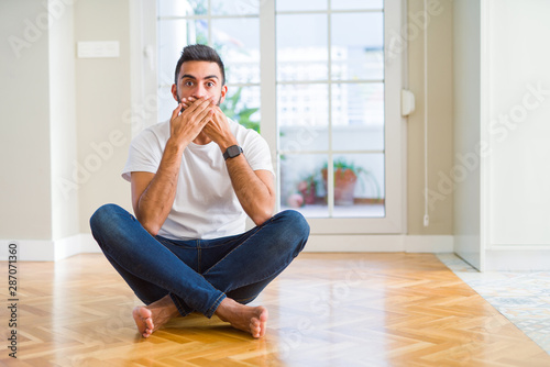 Handsome hispanic man wearing casual t-shirt sitting on the floor at home shocked covering mouth with hands for mistake. Secret concept.