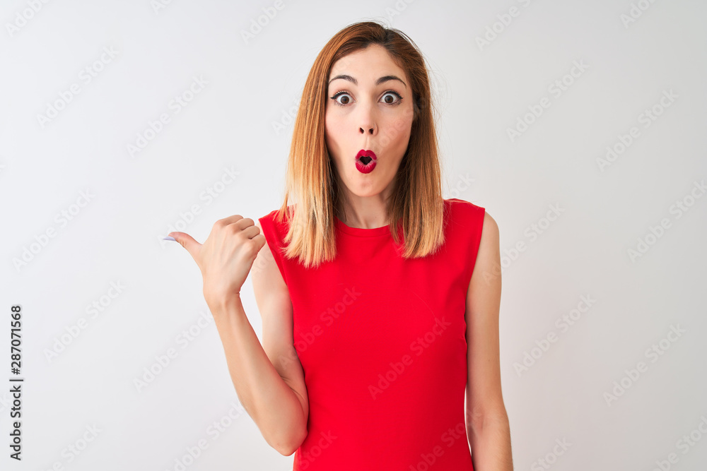 Redhead businesswoman wearing elegant red dress standing over isolated white background Surprised pointing with hand finger to the side, open mouth amazed expression. Redhead businesswoman wearing elegant red dress standing over isolated white background Surprised pointing with hand finger to the side, open mouth amazed expression.