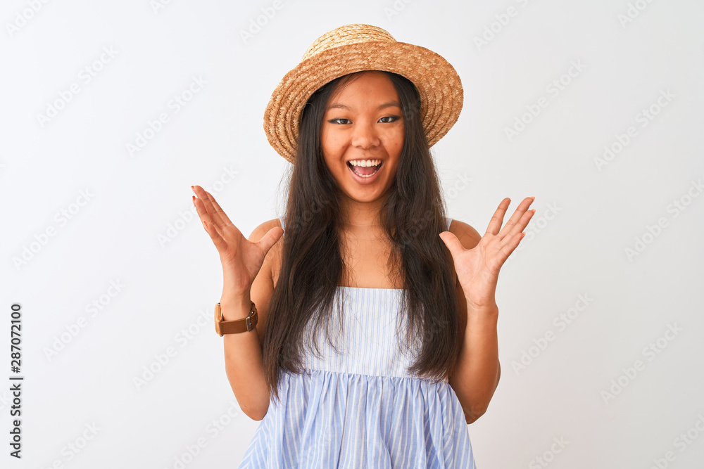 Young chinese woman wearing striped dress and hat standing over isolated white background celebrating crazy and amazed for success with arms raised and open eyes screaming excited. Winner concept Young chinese woman wearing striped dress and hat standing over isolated white background celebrating crazy and amazed for success with arms raised and open eyes screaming excited. Winner concept