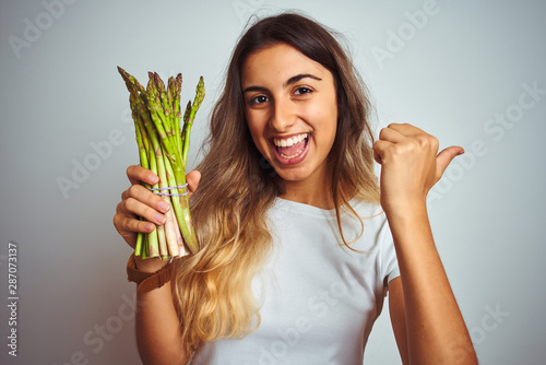 Young beautiful woman eating asparagus over grey isolated background pointing and showing with thumb up to the side with happy face smiling