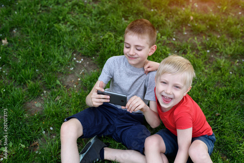 Two boys with a smartphone in their hands are sitting on the grass. © somemeans