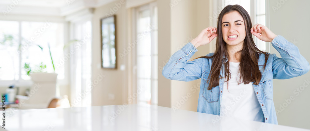 Wide angle picture of beautiful young woman sitting on white table at home covering ears with fingers with annoyed expression for the noise of loud music. Deaf concept. Wide angle picture of beautiful young woman sitting on white table at home covering ears with fingers with annoyed expression for the noise of loud music. Deaf concept.