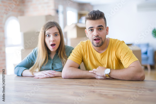 Young couple sitting on the table movinto to new home with carboard boxes behind them afraid and shocked with surprise expression  fear and excited face.