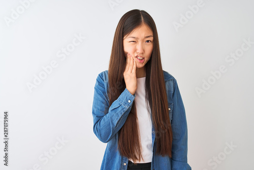 Young chinese woman wearing denim shirt standing over isolated white background touching mouth with hand with painful expression because of toothache or dental illness on teeth. Dentist concept.