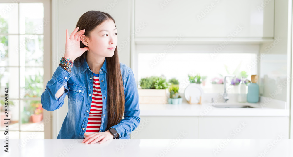 Beautiful Asian woman wearing denim jacket on white table smiling with hand over ear listening an hearing to rumor or gossip. Deafness concept. Beautiful Asian woman wearing denim jacket on white table smiling with hand over ear listening an hearing to rumor or gossip. Deafness concept.