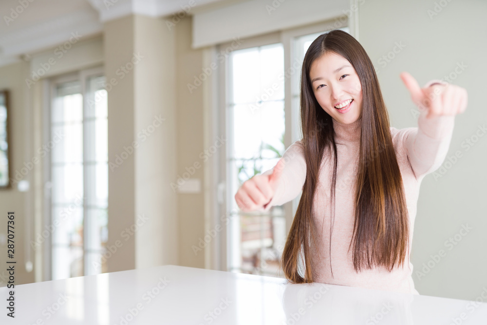 Beautiful Asian woman wearing casual sweater on white table approving doing positive gesture with hand, thumbs up smiling and happy for success. Looking at the camera, winner gesture.