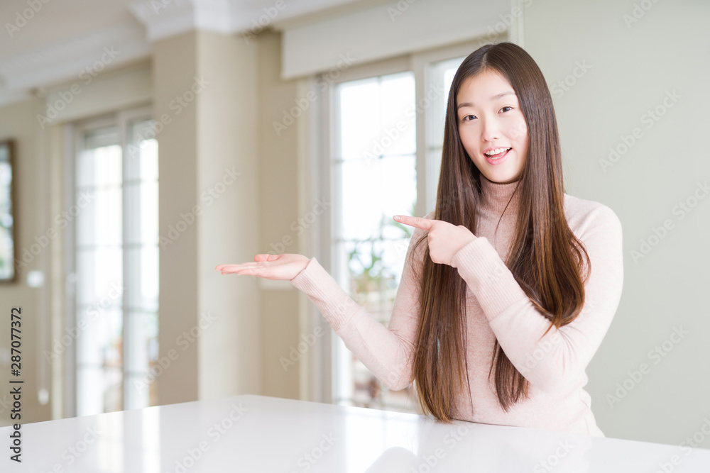Beautiful Asian woman wearing casual sweater on white table amazed and smiling to the camera while presenting with hand and pointing with finger. Beautiful Asian woman wearing casual sweater on white table amazed and smiling to the camera while presenting with hand and pointing with finger.