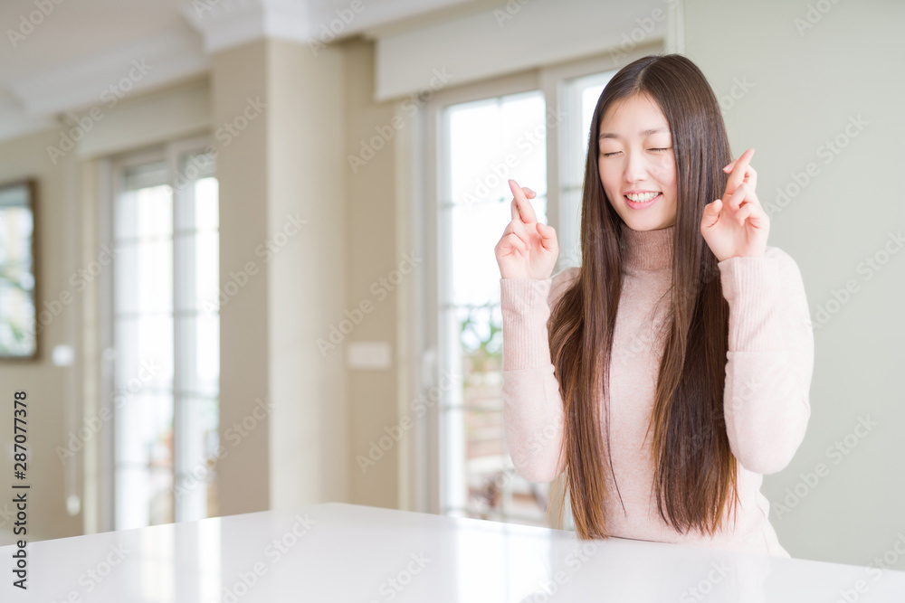 Beautiful Asian woman wearing casual sweater on white table smiling crossing fingers with hope and eyes closed. Luck and superstitious concept. Beautiful Asian woman wearing casual sweater on white table smiling crossing fingers with hope and eyes closed. Luck and superstitious concept.