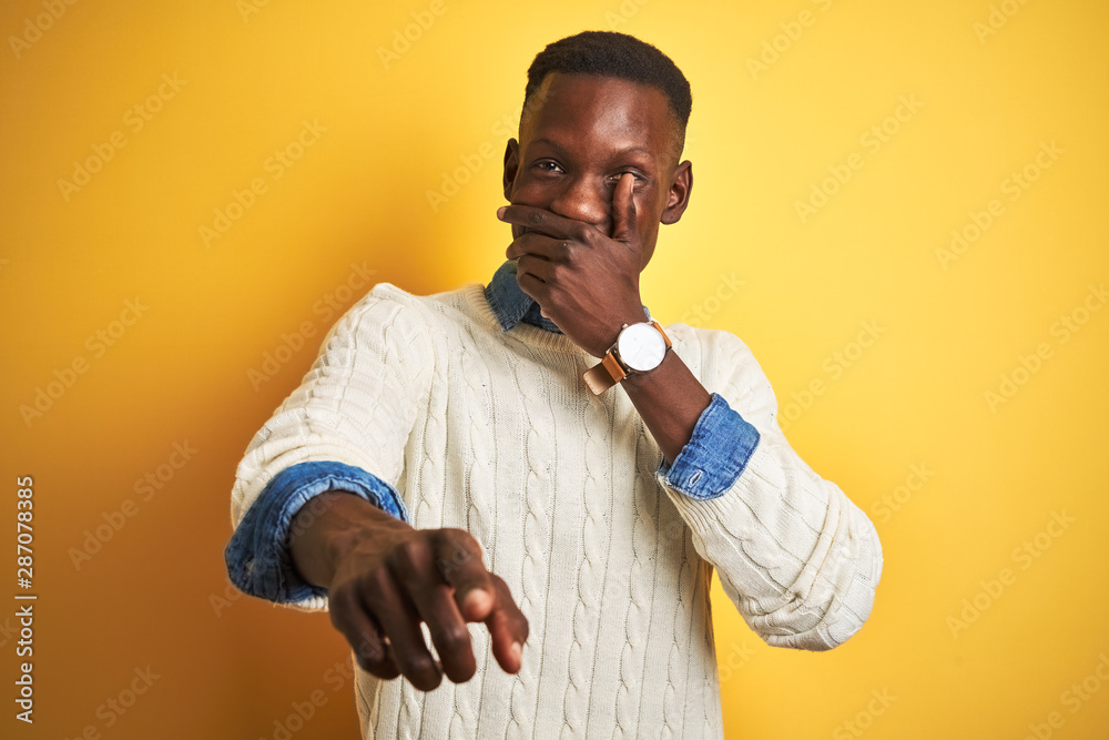 African american man wearing denim shirt and white sweater over isolated yellow background laughing at you, pointing finger to the camera with hand over mouth, shame expression African american man wearing denim shirt and white sweater over isolated yellow background laughing at you, pointing finger to the camera with hand over mouth, shame expression