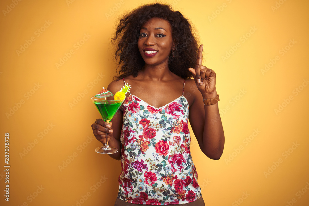 Young african american woman drinking cocktail standing over isolated yellow background surprised with an idea or question pointing finger with happy face, number one